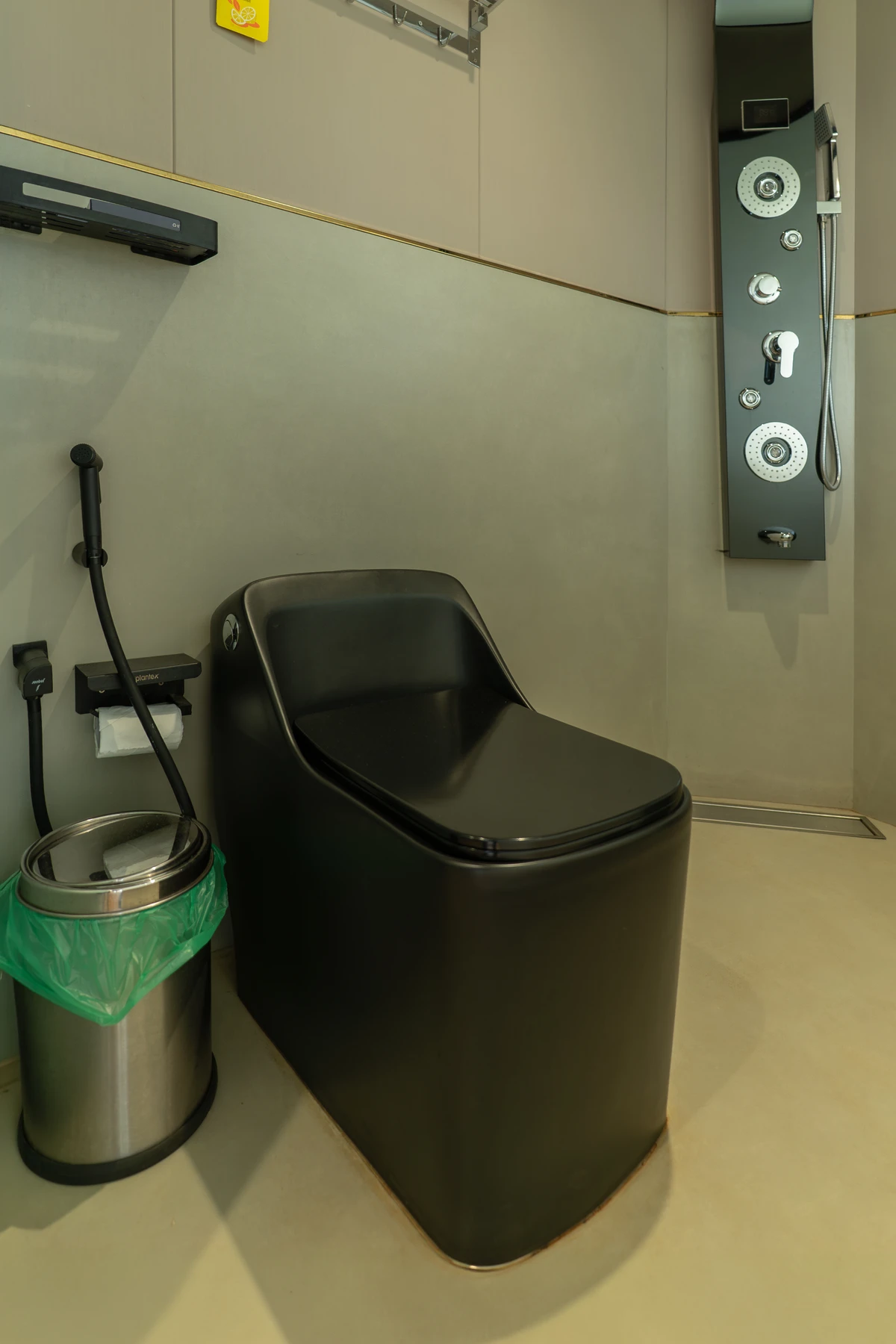 Minimalist Bathroom Stall A modern bathroom stall featuring a matte black toilet, a stainless steel trash can, and a black multi-function shower panel against minimalist beige walls.
